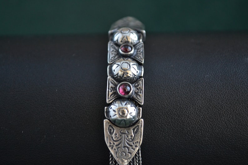 May include: A silver bracelet with a chain design and a series of oval-shaped links. Each link has a raised floral pattern and a red gemstone in the center.