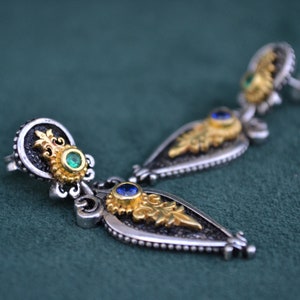 Può includere: Un paio di orecchini in argento e oro con pietre preziose blu e verdi. Gli orecchini hanno una forma a goccia e sono decorati con dettagli intricati.