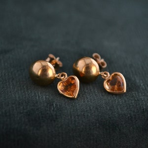 Puede incluir: Un par de pendientes dorados. Cada pendiente presenta un perno redondo y un dije en forma de coraz&oacute;n con una piedra de color &aacute;mbar. Los pendientes se muestran sobre un fondo verde oscuro.