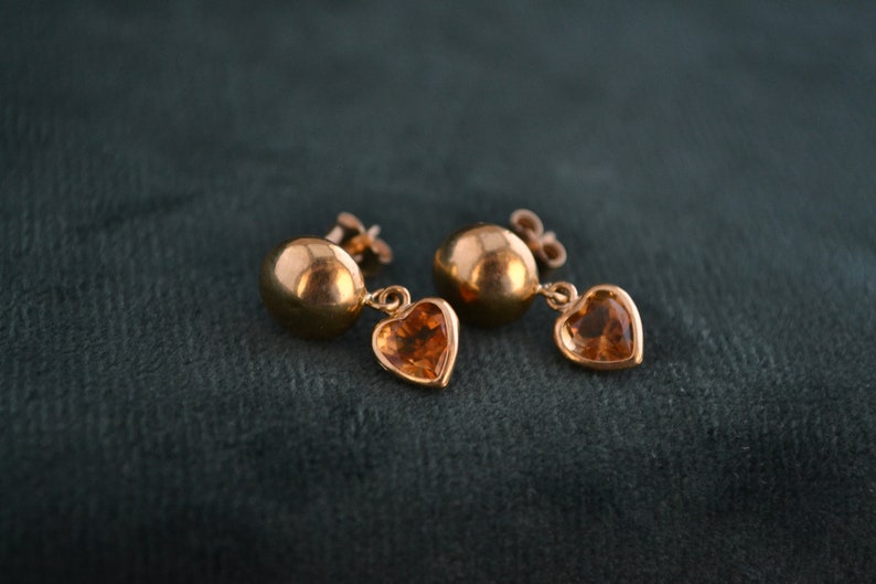 Puede incluir: Un par de pendientes dorados. Cada pendiente presenta un perno redondo y un dije en forma de coraz&oacute;n con una piedra de color &aacute;mbar. Los pendientes se muestran sobre un fondo verde oscuro.