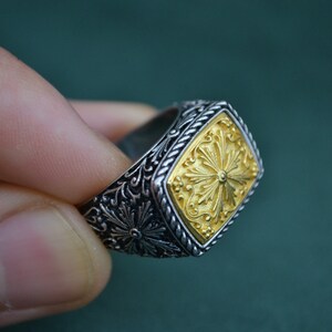 May include: A silver ring with a gold square-shaped design featuring a floral pattern. The ring has a detailed, intricate design.