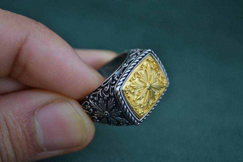 May include: A silver ring with a gold square-shaped design featuring a floral pattern. The ring has a detailed, intricate design.