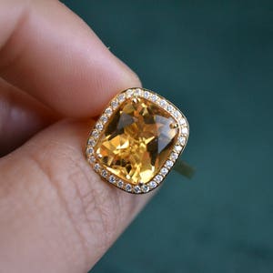Pode incluir: Um anel de ouro com uma grande gema amarela de corte almofada, cercada por um halo de gemas menores e transparentes.