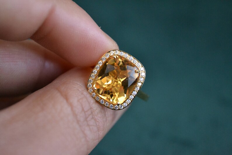 Pode incluir: Um anel de ouro com uma grande gema amarela de corte almofada, cercada por um halo de gemas menores e transparentes.