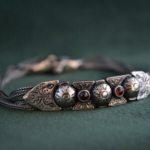 May include: A silver bracelet with a braided chain and five decorative charms. The charms are silver with intricate designs and feature red gemstones.