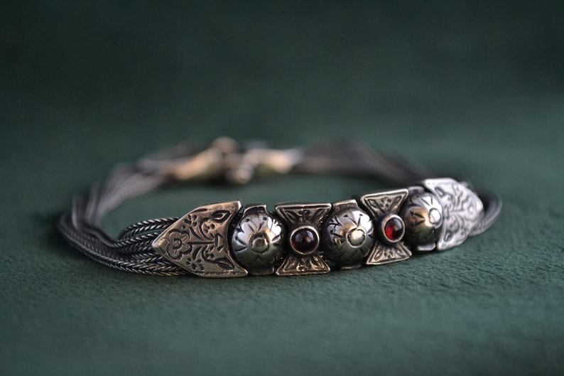 May include: A silver bracelet with a braided chain and five decorative charms. The charms are silver with intricate designs and feature red gemstones.