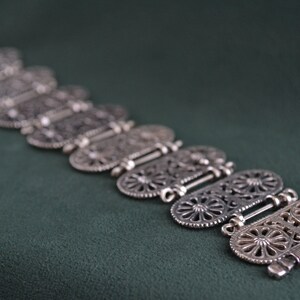 Puede incluir: Un brazalete plateado con un intrincado dise&ntilde;o calado. El brazalete est&aacute; compuesto por varios eslabones ovalados, cada uno con un detallado patr&oacute;n floral o geom&eacute;trico. Los eslabones est&aacute;n conectados por bisagras, sobre una superficie verde oscuro.