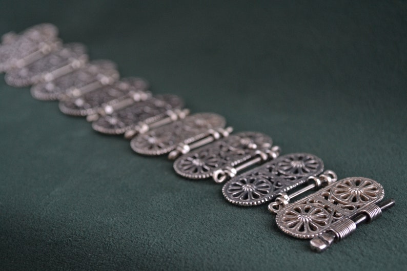 Puede incluir: Un brazalete plateado con un intrincado dise&ntilde;o calado. El brazalete est&aacute; compuesto por varios eslabones ovalados, cada uno con un detallado patr&oacute;n floral o geom&eacute;trico. Los eslabones est&aacute;n conectados por bisagras, sobre una superficie verde oscuro.