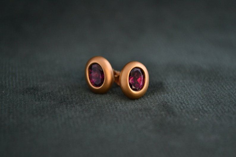 Puede incluir: Un par de pendientes de bot&oacute;n de oro con una piedra ovalada de color rojo oscuro en cada uno.