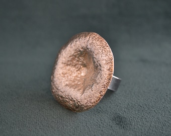 May include: A large, copper-coloured ring with a textured, hammered surface. The ring is shaped like a wide, shallow bowl.