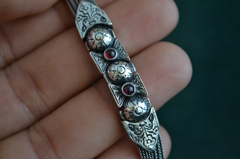 May include: A silver chain bracelet with intricate floral designs and three red gemstones.