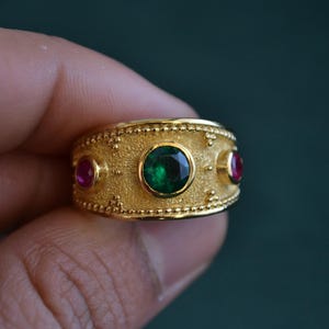 May include: A gold ring with a green gemstone and two smaller red gemstones. The ring has a textured surface and a beaded edge.