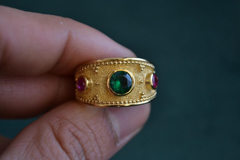May include: A gold ring with a green gemstone and two smaller red gemstones. The ring has a textured surface and a beaded edge.