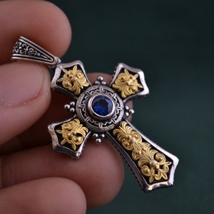 May include: A silver and gold cross pendant with a blue gemstone in the centre. The cross is intricately detailed with floral designs.