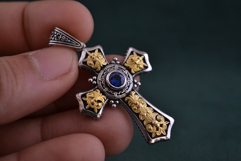 May include: A silver and gold cross pendant with a blue gemstone in the centre. The cross is intricately detailed with floral designs.