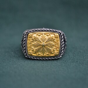 May include: A silver ring with a gold square-shaped center featuring a detailed floral design.
