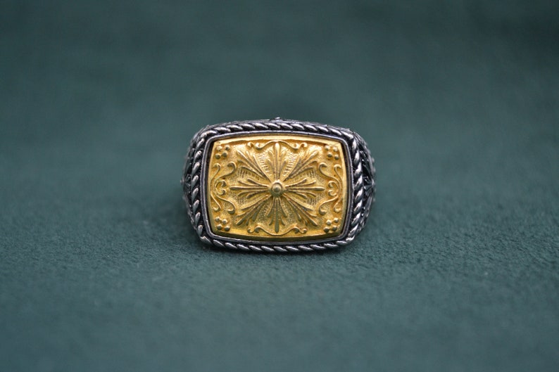 May include: A silver ring with a gold square-shaped center featuring a detailed floral design.