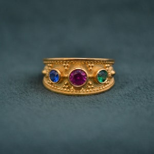 Op de afbeelding: Een gouden ring met drie edelstenen: een blauwe saffier, een roze robijn en een groene smaragd. De ring heeft een getextureerde band.