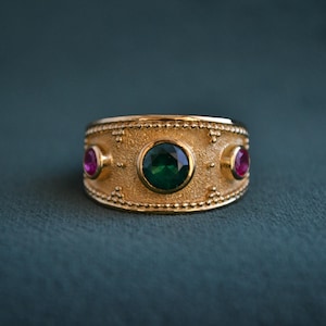May include: A gold ring with a green gemstone and two smaller pink gemstones. The ring has a textured band with a geometric design.