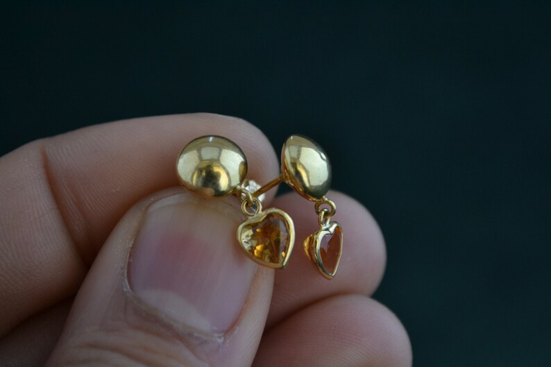Puede incluir: Pendientes de bot&oacute;n dorados con colgantes en forma de coraz&oacute;n. Los pendientes presentan un bot&oacute;n redondo y una peque&ntilde;a gema en forma de coraz&oacute;n. Las gemas son de color &aacute;mbar c&aacute;lido. Los pendientes se sostienen.
