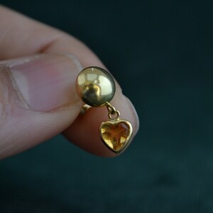 Puede incluir: Un pendiente de color dorado con un peque&ntilde;o colgante en forma de coraz&oacute;n de piedra preciosa de color &aacute;mbar. El pendiente tiene una parte superior redonda y se sostiene en la mano de una persona sobre un fondo verde oscuro.