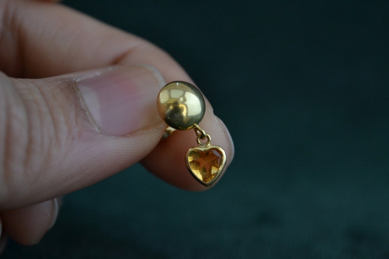 Puede incluir: Un pendiente de color dorado con un peque&ntilde;o colgante en forma de coraz&oacute;n de piedra preciosa de color &aacute;mbar. El pendiente tiene una parte superior redonda y se sostiene en la mano de una persona sobre un fondo verde oscuro.