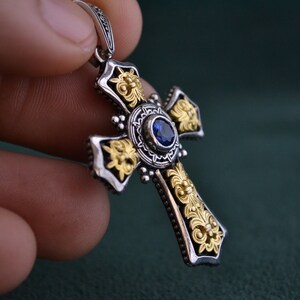 May include: A silver and gold cross pendant with a blue gemstone in the centre. The cross is decorated with intricate floral designs.