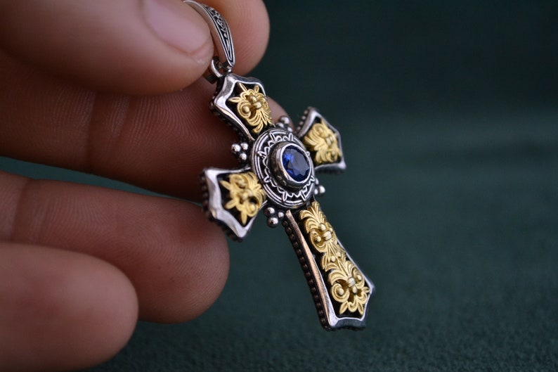 May include: A silver and gold cross pendant with a blue gemstone in the centre. The cross is decorated with intricate floral designs.