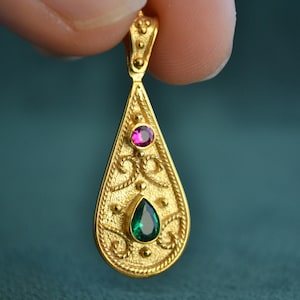 Pode incluir: Um pingente de ouro em forma de lágrima, com intrincados desenhos florais e duas pedras preciosas. A pedra preciosa superior é rosa e a pedra preciosa inferior é verde.