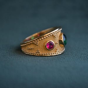 May include: A gold ring with a textured band and two gemstones. The larger gemstone is green and the smaller gemstone is red.