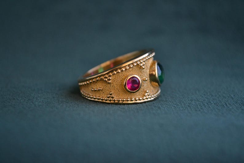 May include: A gold ring with a textured band and two gemstones. The larger gemstone is green and the smaller gemstone is red.