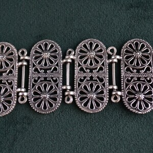 Puede incluir: Pulsera plateada con un intrincado dise&ntilde;o floral. La pulsera est&aacute; compuesta por m&uacute;ltiples eslabones ovalados, cada uno adornado con un detallado patr&oacute;n de flores. Los eslabones est&aacute;n conectados por barras decorativas m&aacute;s peque&ntilde;as y se cierran con un cierre seguro.