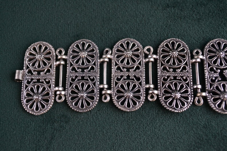 Puede incluir: Pulsera plateada con un intrincado dise&ntilde;o floral. La pulsera est&aacute; compuesta por m&uacute;ltiples eslabones ovalados, cada uno adornado con un detallado patr&oacute;n de flores. Los eslabones est&aacute;n conectados por barras decorativas m&aacute;s peque&ntilde;as y se cierran con un cierre seguro.
