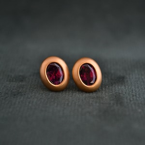 Puede incluir: Un par de pendientes ovalados con un acabado en oro rosa y una piedra preciosa roja oscura en el centro de cada pendiente.