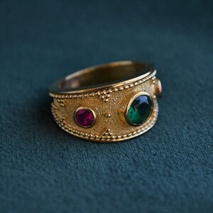 May include: A gold ring with a textured band and three gemstones. The ring has a green gemstone in the center and two smaller pink gemstones on either side.