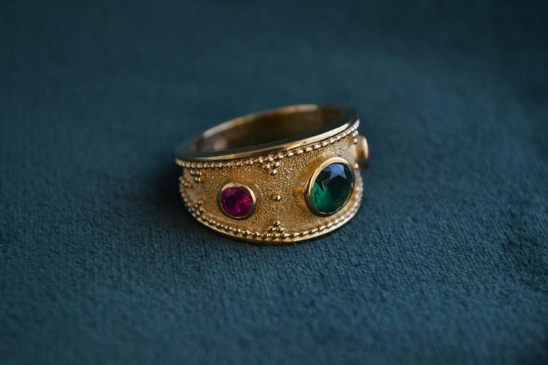 May include: A gold ring with a textured band and three gemstones. The ring has a green gemstone in the center and two smaller pink gemstones on either side.