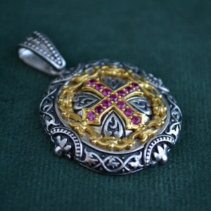 Könnte beinhalten: Ein silberner und goldener Anhänger mit einem roten, mit Edelsteinen besetzten Kreuz in der Mitte. Der Anhänger ist von einem komplizierten Design aus Silber und Gold umgeben.