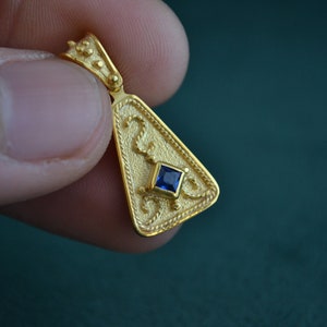 May include: A gold pendant shaped like a triangle, featuring intricate detailing and a small blue gemstone at its centre.