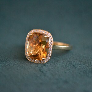 Pode incluir: Um anel de ouro com uma grande pedra preciosa laranja em forma de quadrado, um top&aacute;zio, rodeado por um halo de pequenas pedras preciosas transparentes.