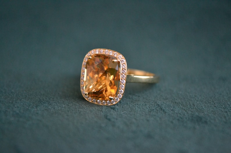 Pode incluir: Um anel de ouro com uma grande pedra preciosa laranja em forma de quadrado, um top&aacute;zio, rodeado por um halo de pequenas pedras preciosas transparentes.