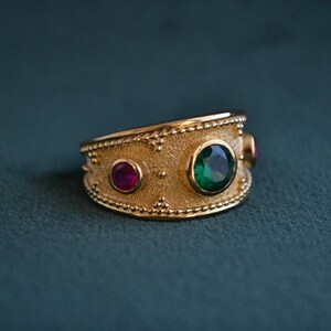 May include: A gold ring with a textured band and three gemstones. The center stone is a large, emerald green gemstone. The two smaller stones are ruby red.
