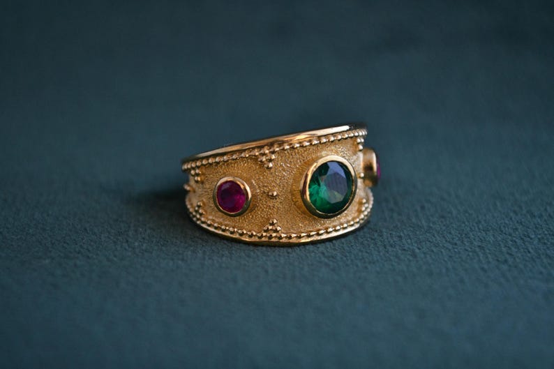 May include: A gold ring with a textured band and three gemstones. The center stone is a large, emerald green gemstone. The two smaller stones are ruby red.