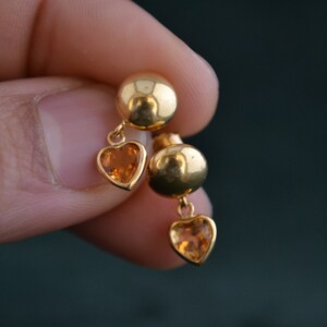 Puede incluir: Un par de pendientes dorados. Cada pendiente presenta un peque&ntilde;o perno redondo y un dije en forma de coraz&oacute;n con una piedra preciosa naranja. Los pendientes se sostienen contra un fondo borroso.