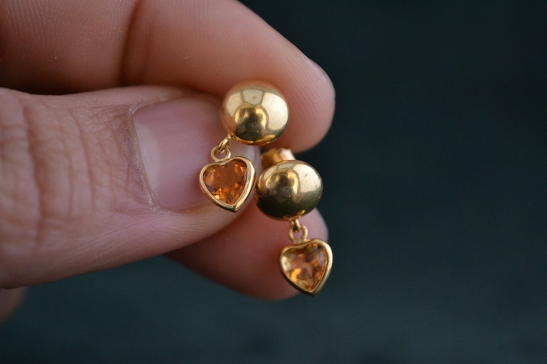 Puede incluir: Un par de pendientes dorados. Cada pendiente presenta un peque&ntilde;o perno redondo y un dije en forma de coraz&oacute;n con una piedra preciosa naranja. Los pendientes se sostienen contra un fondo borroso.