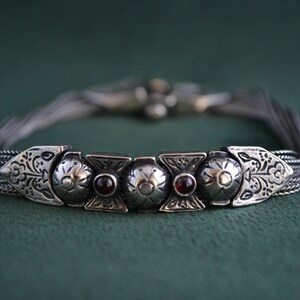 May include: A silver bracelet with a decorative design featuring five round, silver charms with red gemstones. The bracelet is made of multiple strands of silver chain.