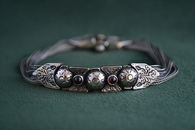 May include: A silver bracelet with a decorative design featuring five round, silver charms with red gemstones. The bracelet is made of multiple strands of silver chain.