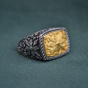 May include: A silver ring with a gold square-shaped center piece. The center piece has a detailed floral design.