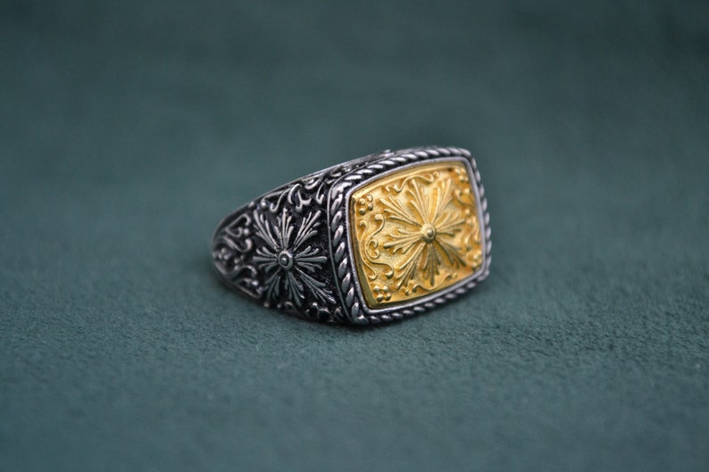 May include: A silver ring with a gold square-shaped center piece. The center piece has a detailed floral design.