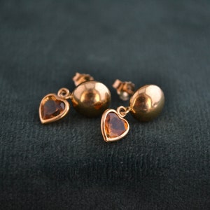 Puede incluir: Un par de pendientes dorados. Cada pendiente presenta una bola redonda de oro pulido y un dije en forma de coraz&oacute;n con una gema marr&oacute;n. Los pendientes est&aacute;n sobre un fondo verde oscuro.