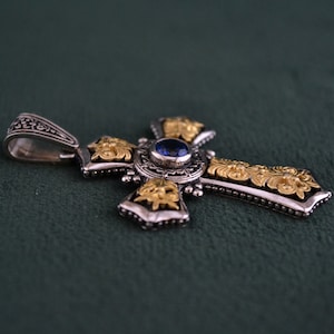 May include: A silver and gold cross pendant with a blue gemstone in the centre. The cross is decorated with intricate floral designs.